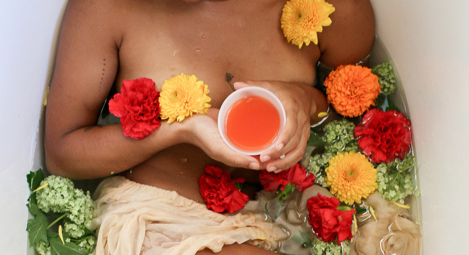 Person in a bathtub with flowers and holding a cup of red liquid