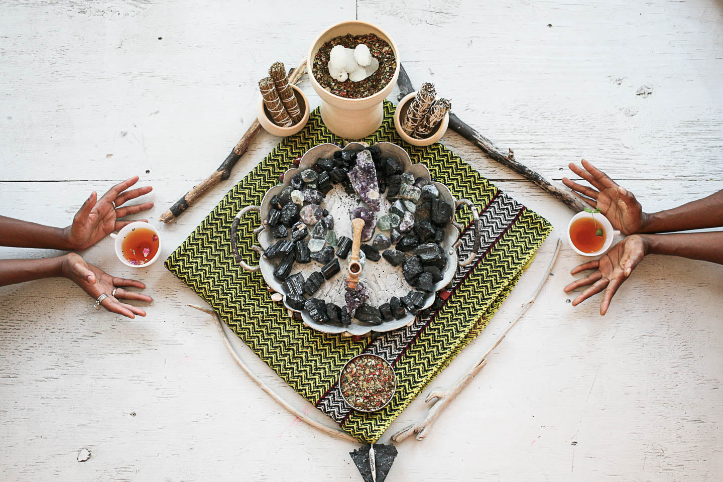 Two people holding cups of tea over a small altar with stones and plants on a white surface.