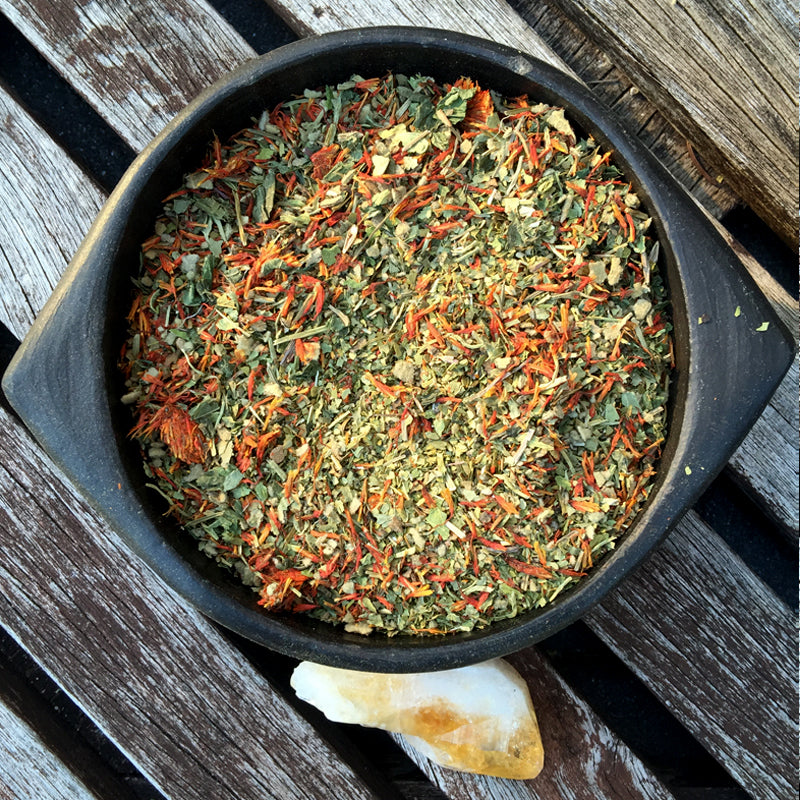 Black ceramic bowl with red and green herbs from AWAKE Herbal Blend next to a yellow crystal on wooden surface