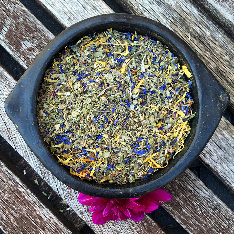 Ceramic black bowl of CENTER Herbal Blend herbs on wood panels next to a fuchsia flower