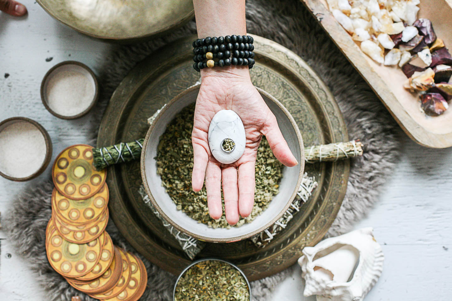 Hand holding a stone over a bowl of herbs with various natural items on a textured surface