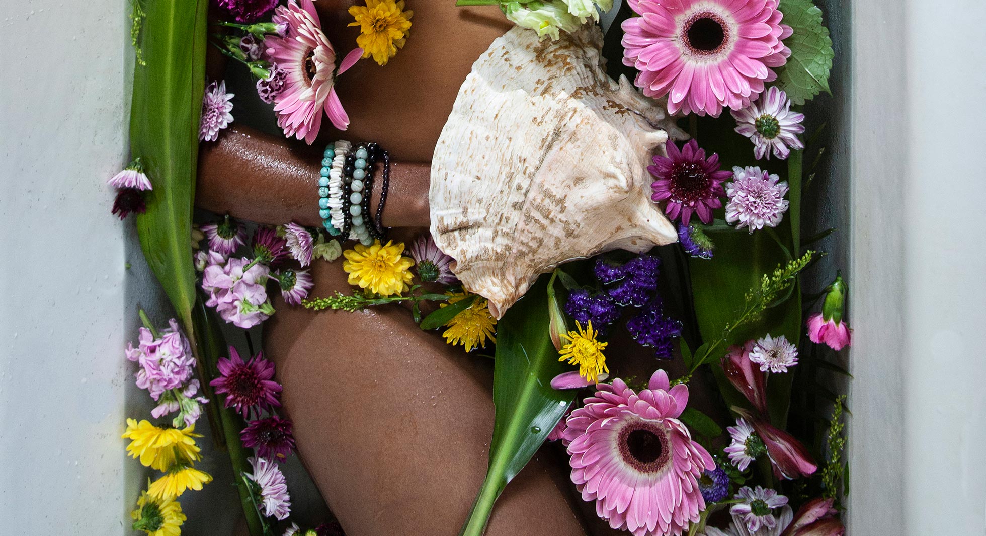 Person wrapped in flowers with a shell, surrounded by vibrant floral arrangement.