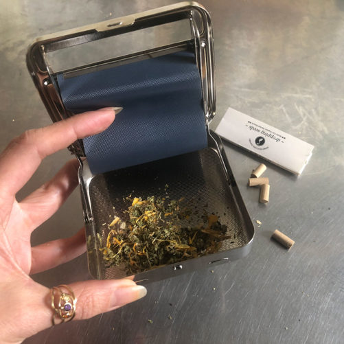 Step 4 Hand holding herbal roller with a mix of herbs and Cannabis next to herbal tin with rolling papers on steel table.