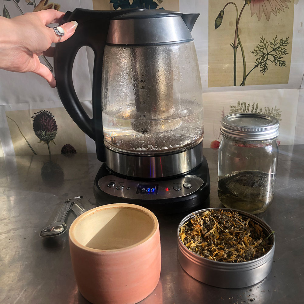 Hand holding electric kettle with pink tea cup and SUN Herbal blend.